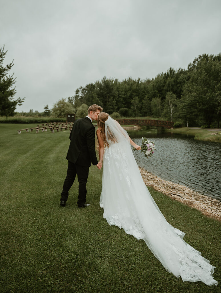 a bride and groom facing away from the camera and kissing along the edge of the pond 