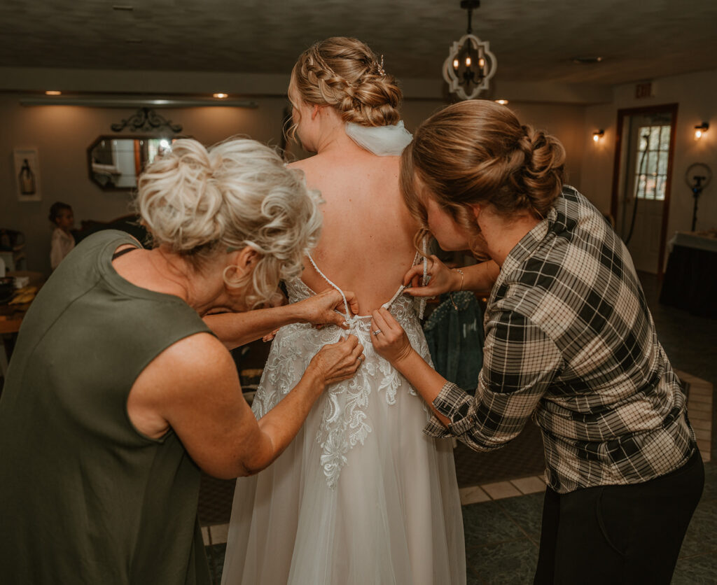 the bride is getting her dress buttoned up by important ladies in her life