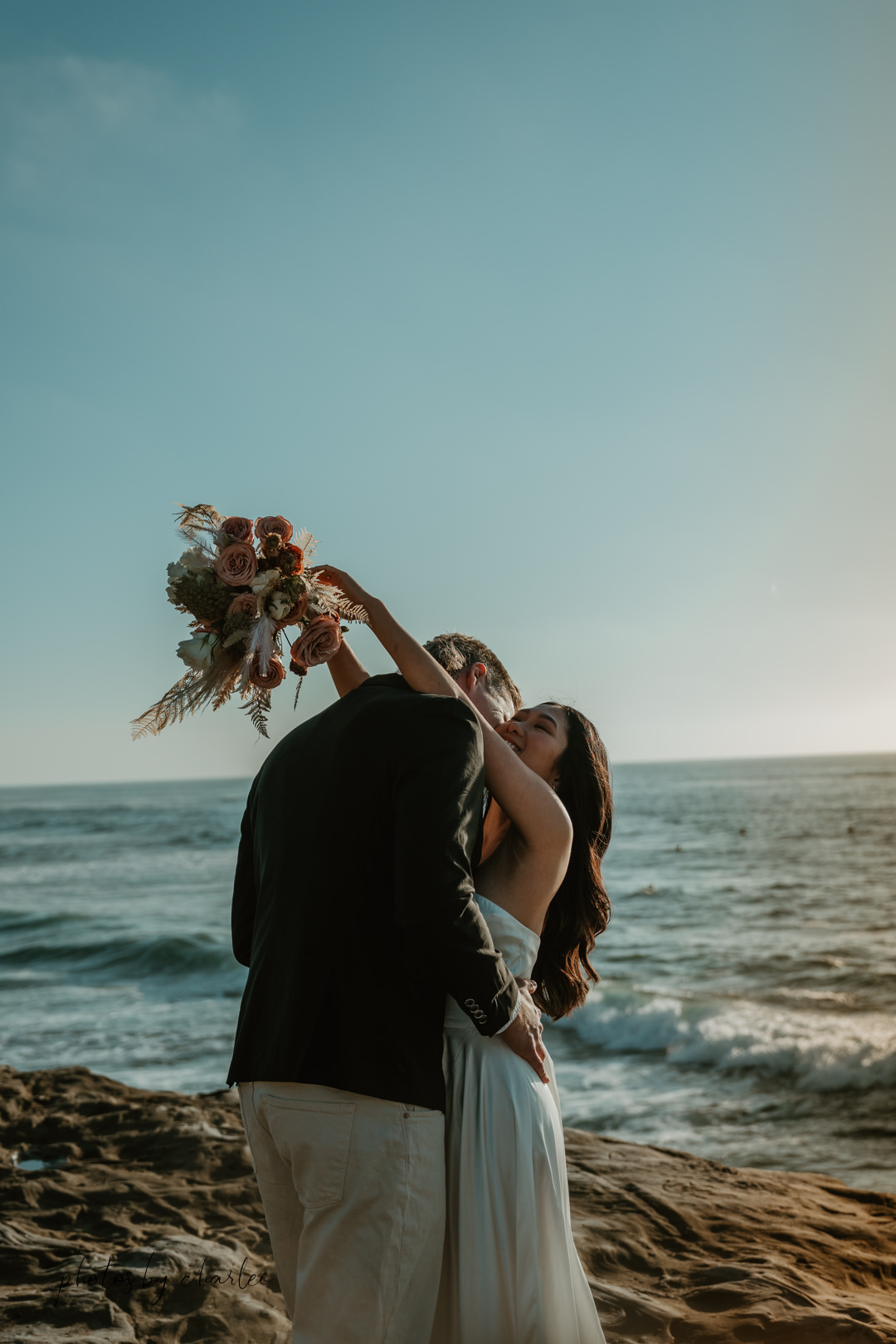 Intimate moment during a elopement: bride wraps her arms around groom's neck, holding a beautiful bouquet, with the San Diego coastline in the background